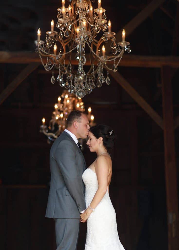 groom kissing brides forhead in barn, chandelier hanging above