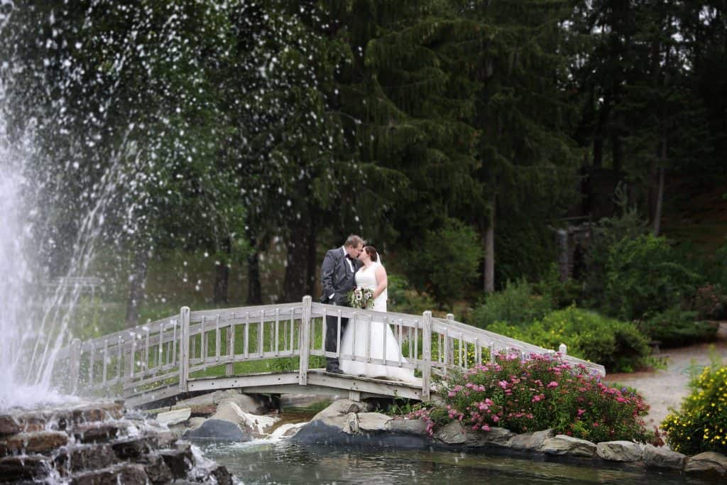 bride and groom on a bridge over water, water fountain in front, green trees in the back