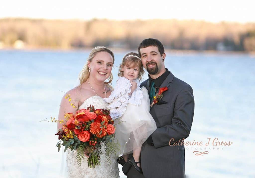 bride and groom with child looking at the camera, lake in background, red and orange flowers