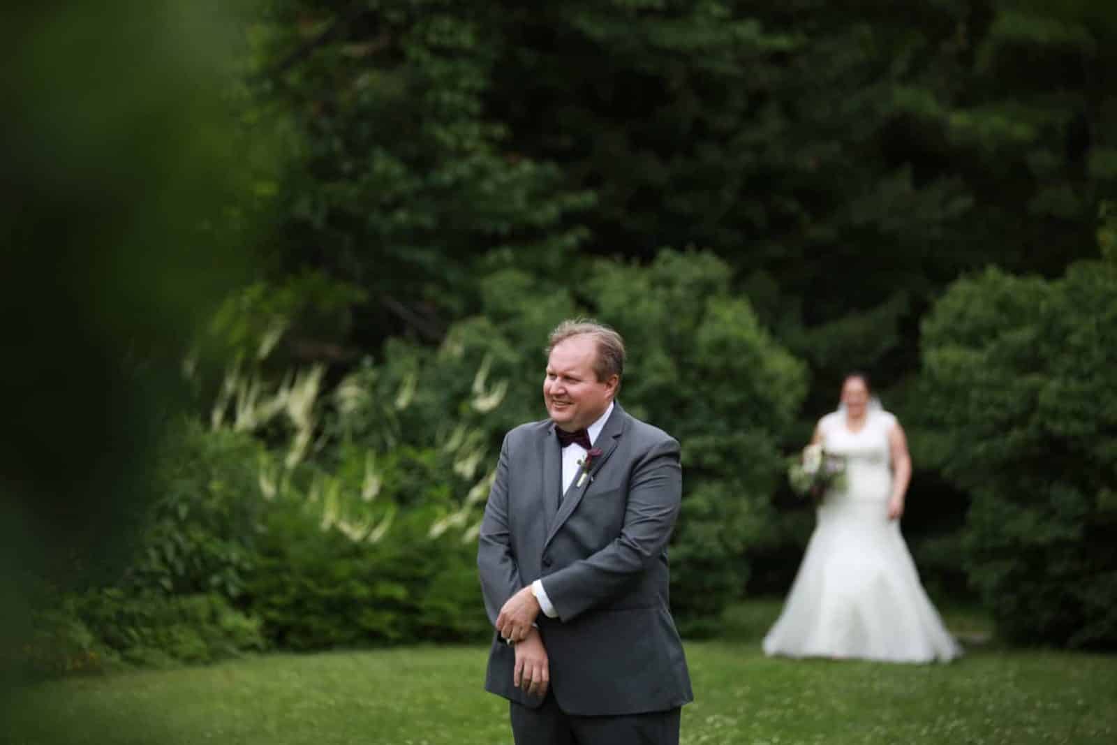 First Look. Groom in focus with a smile bride behind him walking towards grooms back in the ornamental garden at Cascade Park Bangor Maine