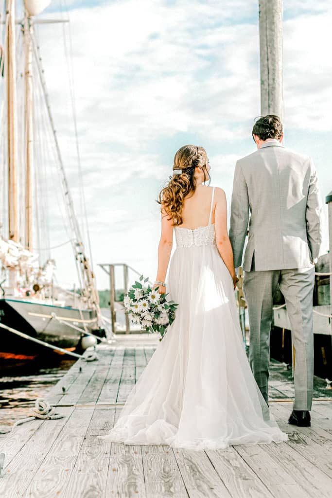 bride and groom walking along a dock next to a sailboat in maine while maine wedding videographer films