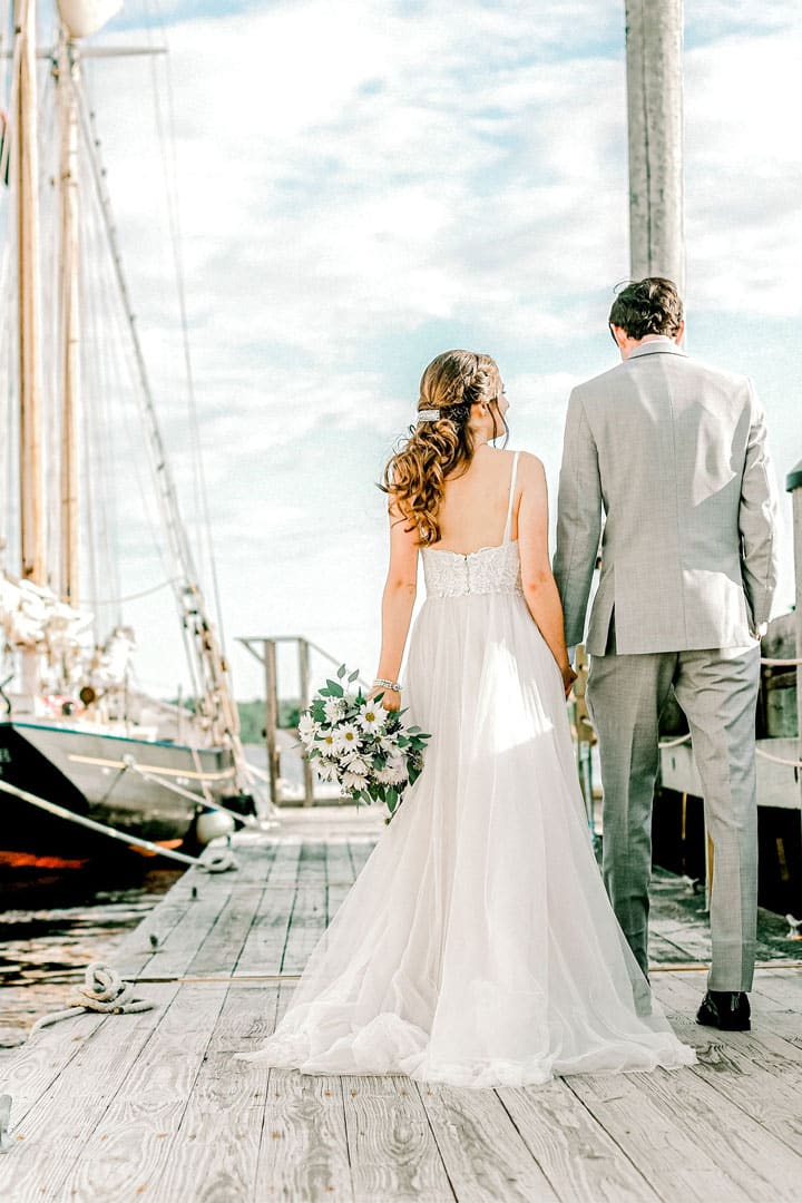 Portland Maine wedding photographer captures bride and groom walking along a dock next to a sailboat in Portland Maine