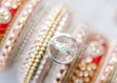 A close-up of a silver ring with a diamond set against a blurred background of intricate, colorful bangles adorned with gold and red embellishments.