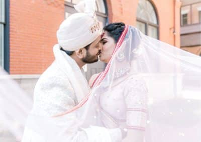 A bride and groom share a kiss under a sheer veil. The groom wears a traditional white outfit and turban, while the bride wears a white and red embellished sari. They stand in front of a red brick building at portland's ocean gateway in Maine
