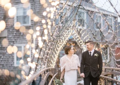 A bride and groom hold hands while walking under an archway covered in twinkling lights. The bride wears a white dress and fur shrug, and the groom is in a black suit. They are smiling at each other against a backdrop of rustic buildings in portland maine