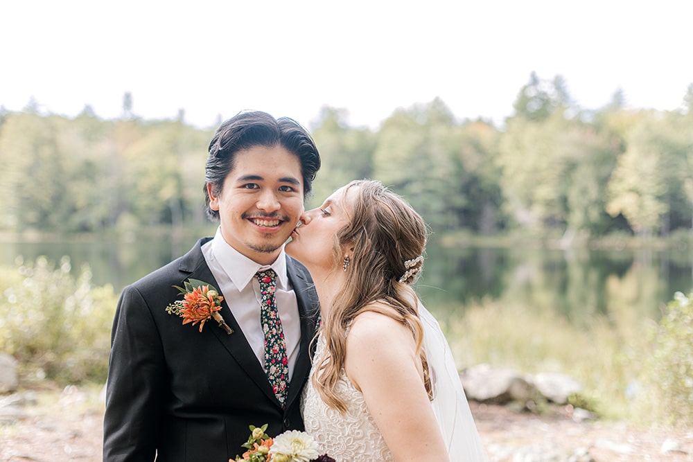 bride kissing grooms cheek with a private Maine lake in the background