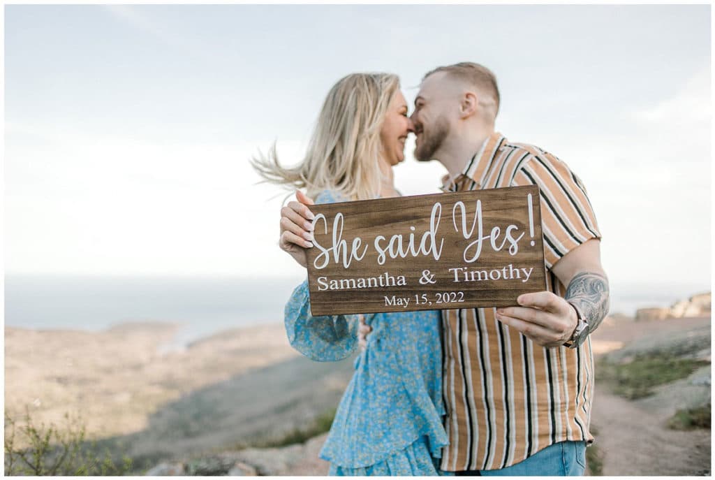 newly engaged couple holding up sign saying she said yes while kissing