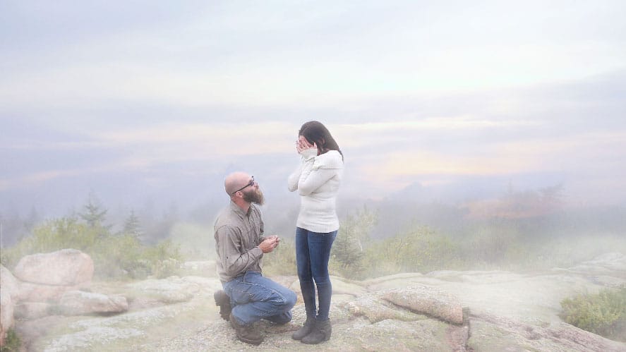 A man kneels on rocky ground, holding a ring box, proposing to a woman with long hair. She covers her face with her hands in surprise and joy. They are outdoors, surrounded by mist and a serene landscape.