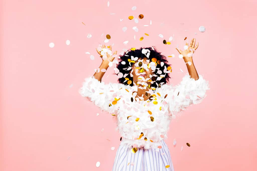 woman celebrating by throwing metallic confetti in front of herself and a pink background