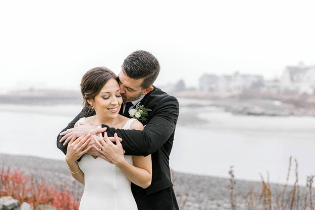 A couple embraces lovingly by a foggy waterfront at breakwater inn and spa in Maine. The man, in a dark suit, wraps his arms around the woman, who is wearing a white dress. They stand on a rocky shoreline with blurred houses in the background.