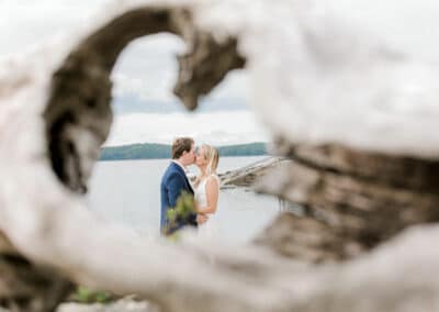 Maine wedding photography by Catherine J. Gross, showcasing A couple shares a kiss on a beach, framed artistically by a hole in a tree trunk. The background features a serene water view with a cloudy sky. The image captures a romantic and intimate moment in nature.