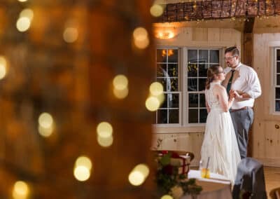 Catherine J. Gross Photography captures A couple dancing in a warmly lit room with large windows. Soft, blurred lights in the foreground create a dreamy atmosphere. The man wears a white shirt and tie, and the woman wears a long dress. A drink sits on a nearby table.