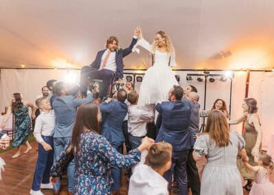 Catherine J. Gross Photography captures a stunning Maine wedding moment showcasing A bride and groom are lifted on chairs by guests at a wedding reception. The bride wears a white dress, and the groom is in a suit. They hold hands joyfully above the crowd. People around them are smiling and celebrating.